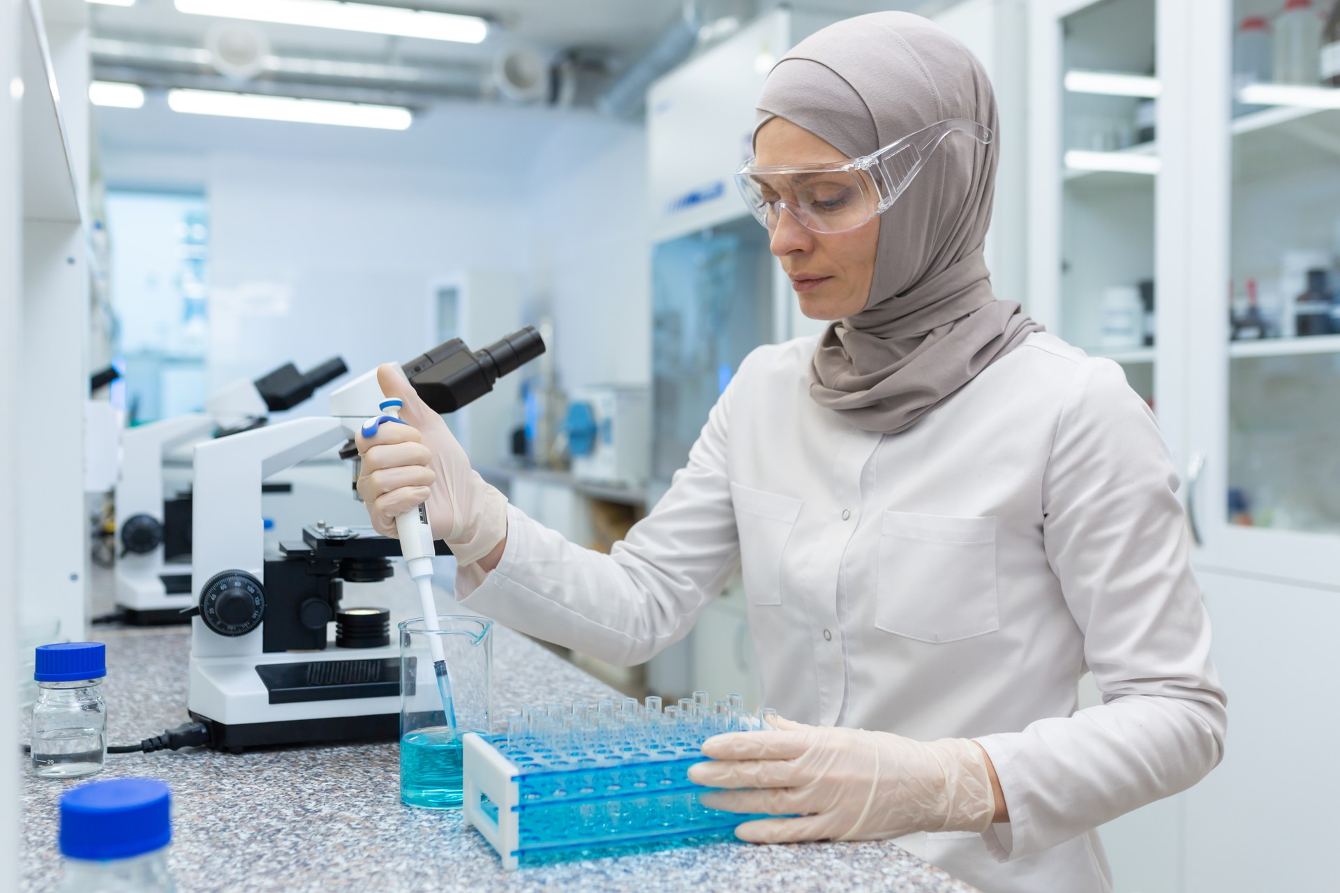 Muslim young woman in hijab, scientist and pharmacist in laboratory. Sitting at table and working with liquids and flasks